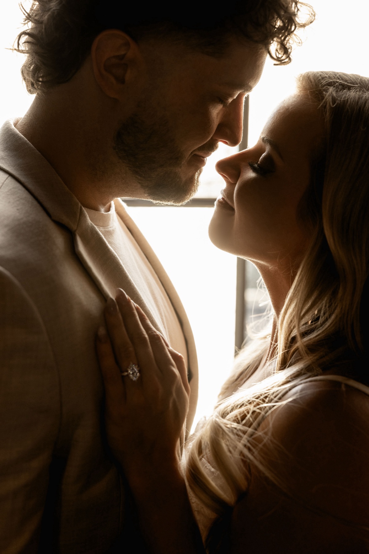Backlit by the window, faces close, her hand on his chest with the ring catching the light.